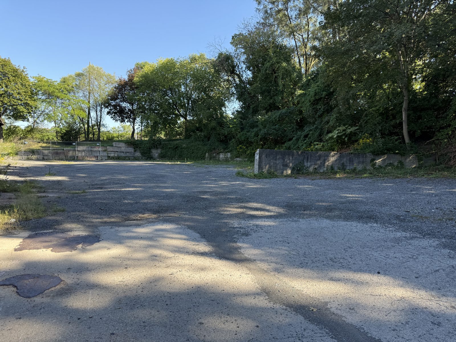 The yard today: a gravel lot with low concrete retaining walls against a line of trees, no structure on it yet