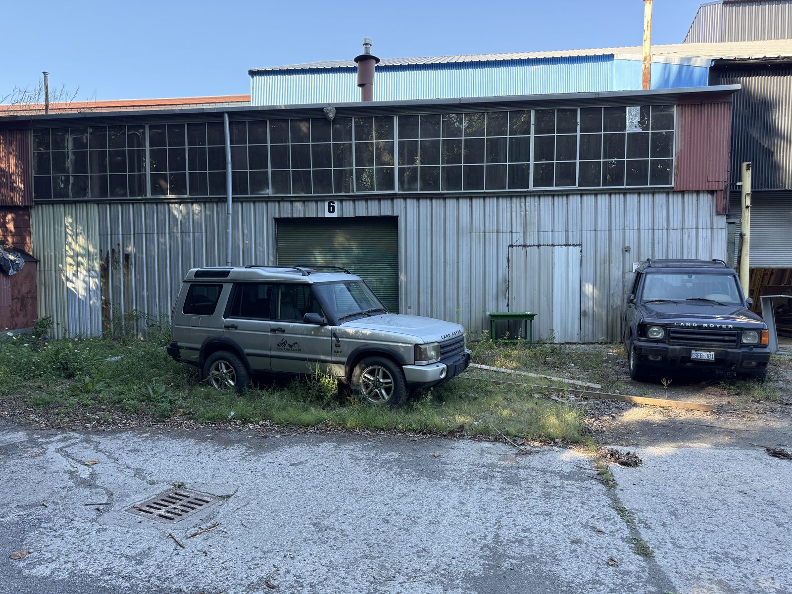 The existing clerestory building today: grey corrugated cladding under a band of windows, with a central doorway and vehicles parked in front