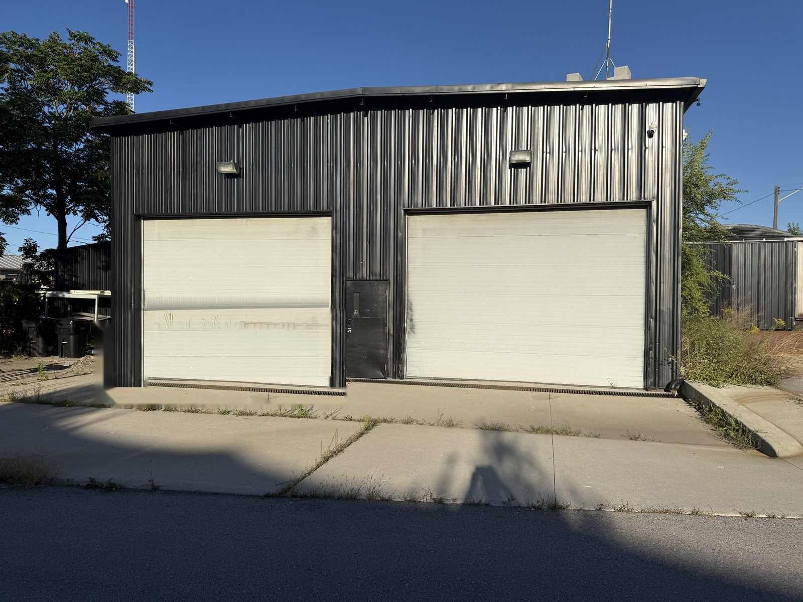The existing two-bay garage today: a black corrugated-steel building with two white roll-up doors and a centre door on a concrete apron