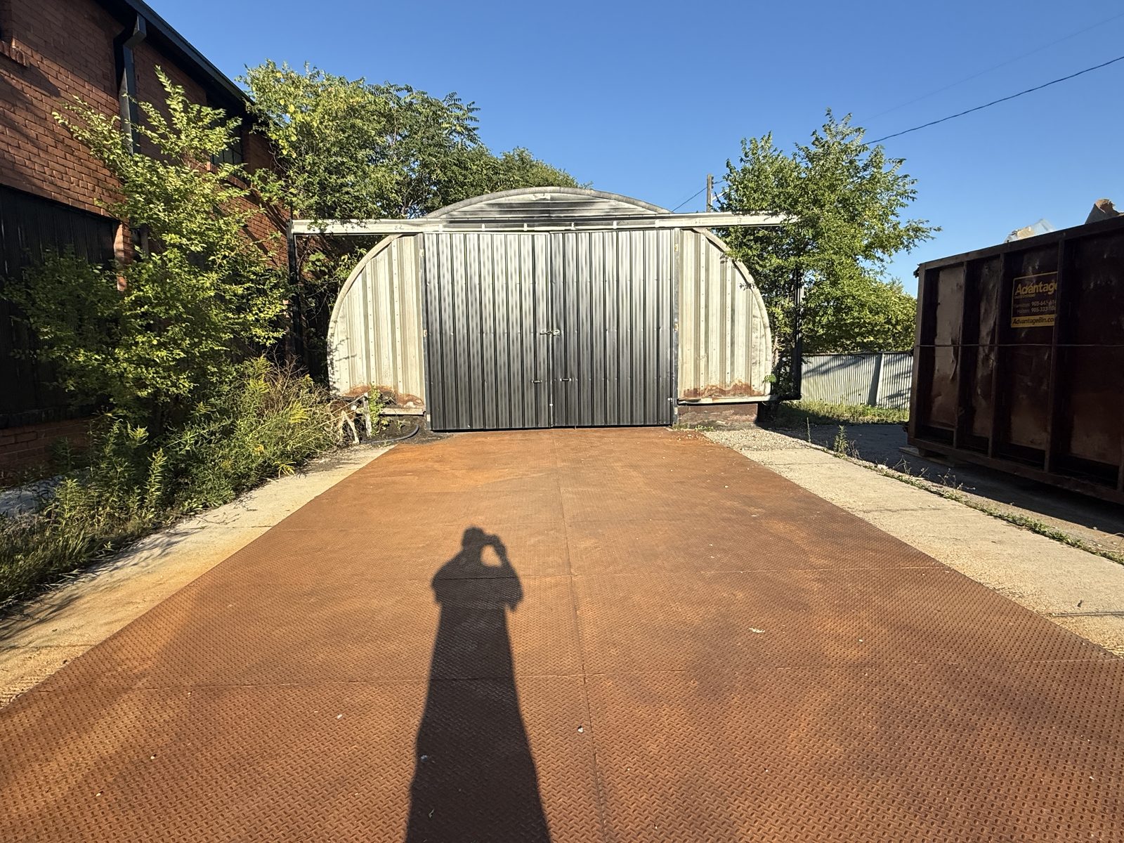 The existing Quonset hut today: an arched corrugated hut at the end of a rusted steel-plate drive, between chain-link fences