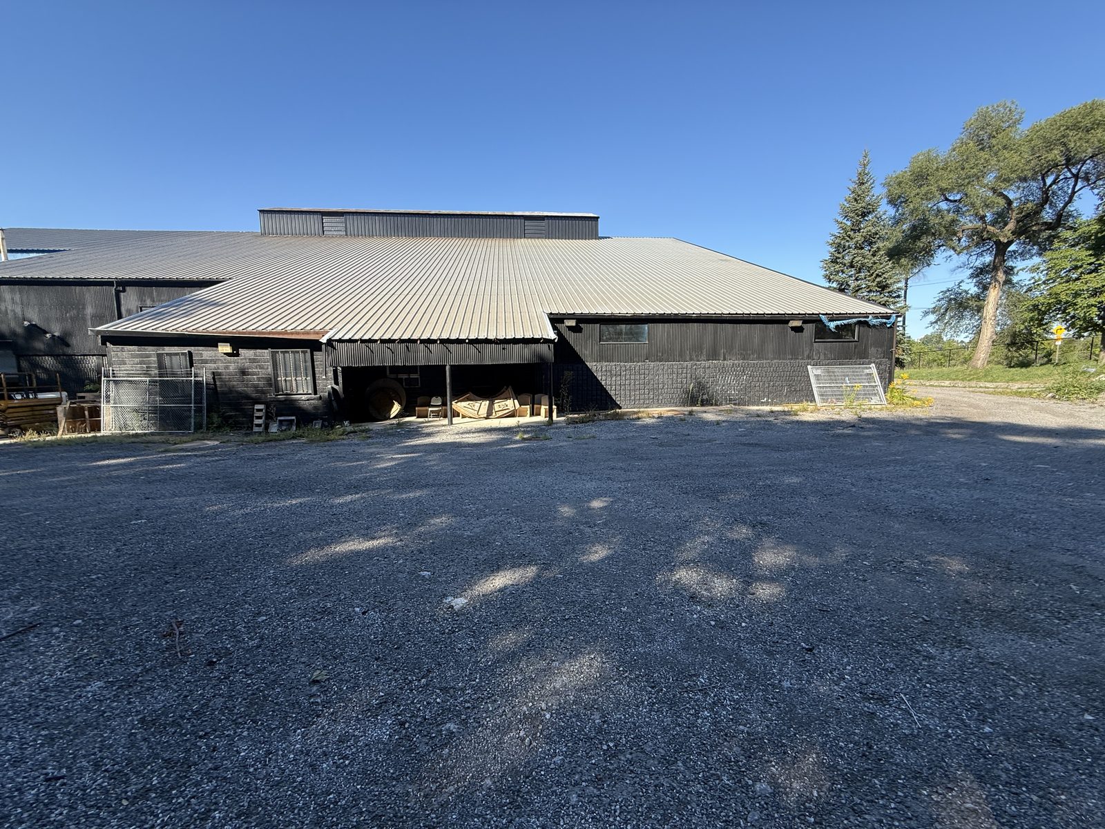The existing building today: a low black building with a wide pitched metal roof and a covered entrance, on a gravel lot with trees behind