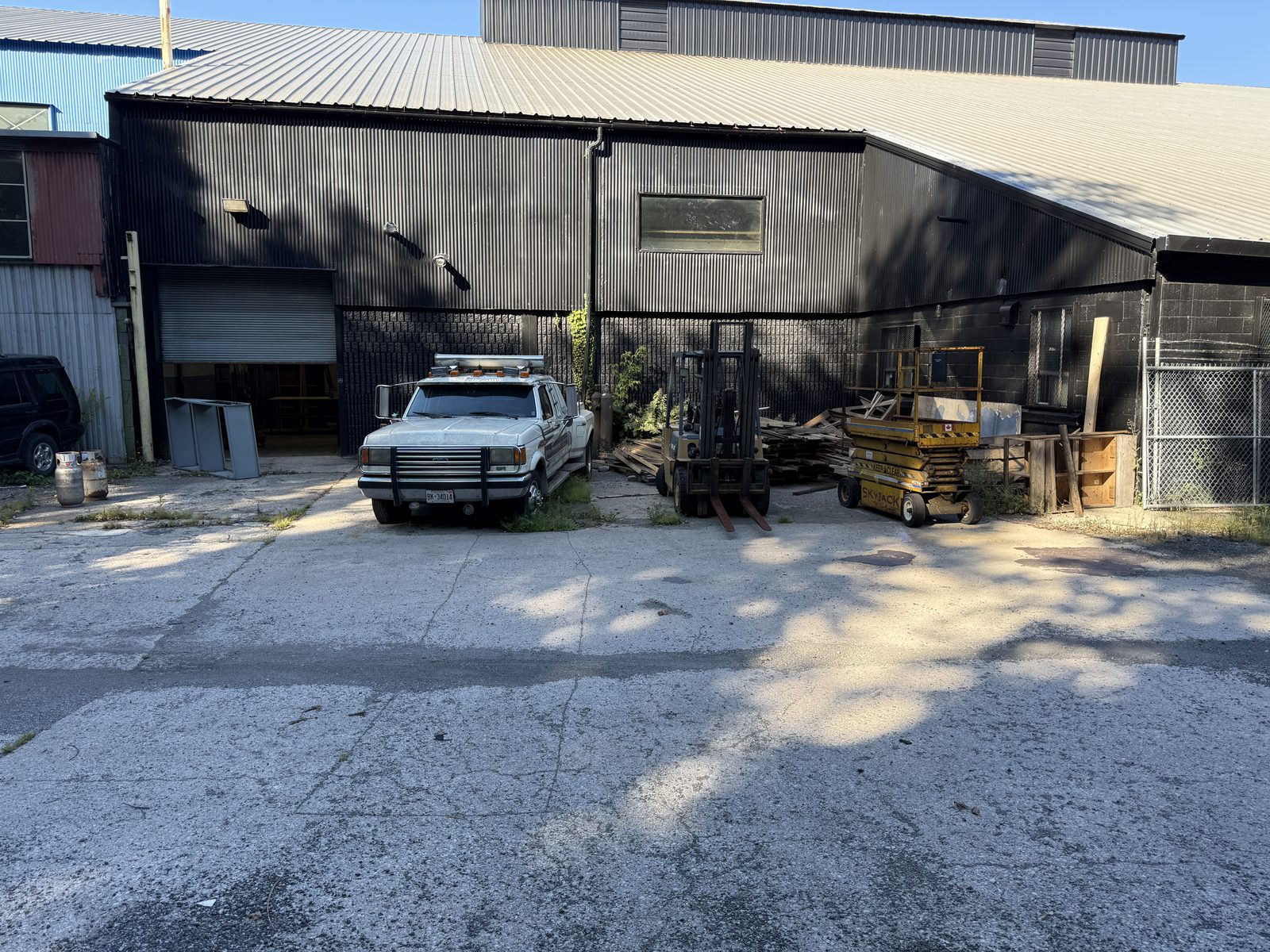 The existing building today: a long dark-clad industrial building with a roller door, a pale metal roof and a concrete yard